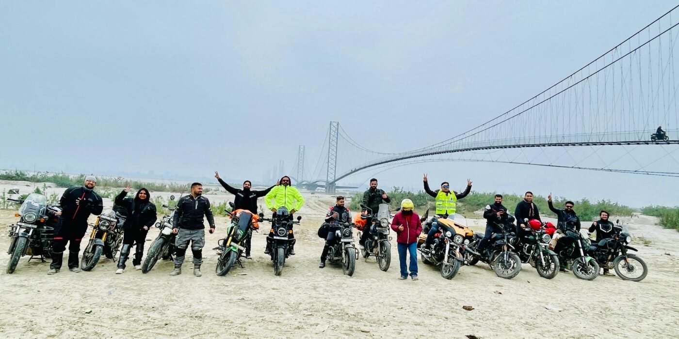 Group of motorcyclists lined up on a riverbed in front of a long suspension bridge on a misty morning