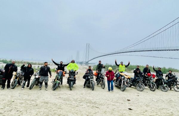 Group of motorcyclists lined up on a riverbed in front of a long suspension bridge on a misty morning