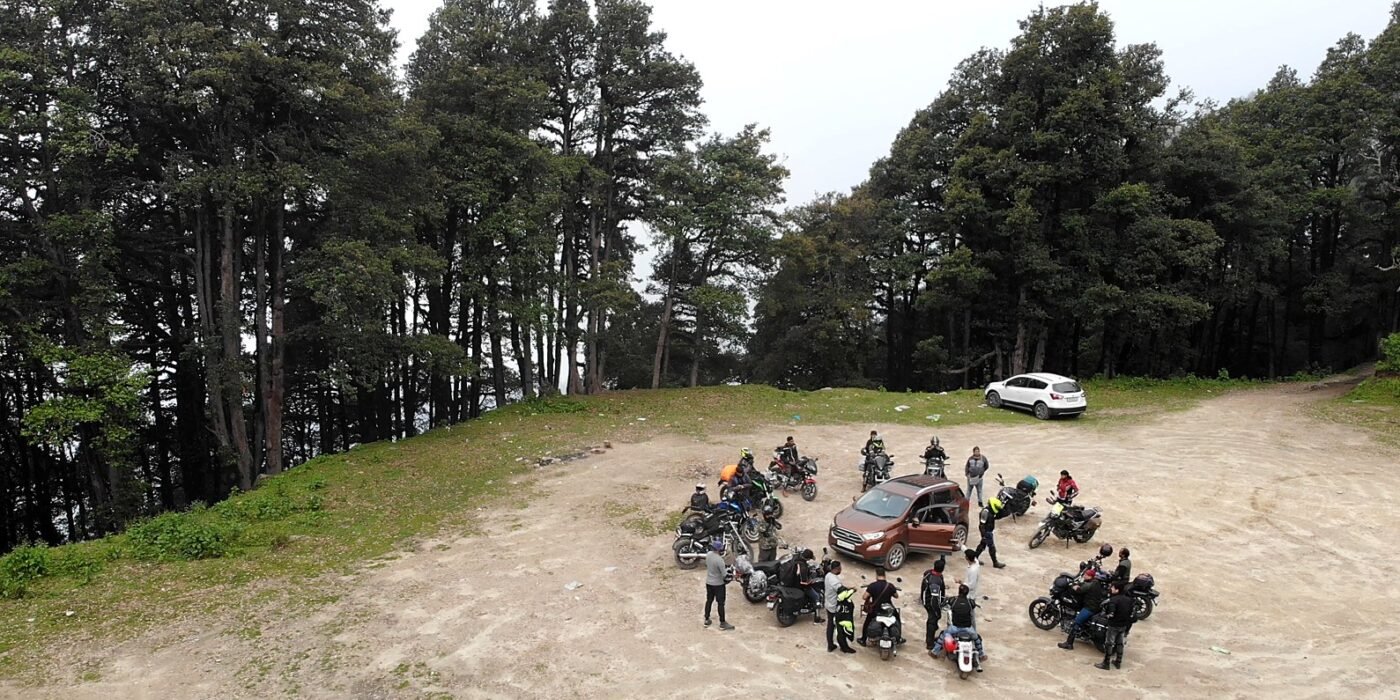 Riders and motorcycles gathered in a forest clearing among tall trees on a weekend getaway ride